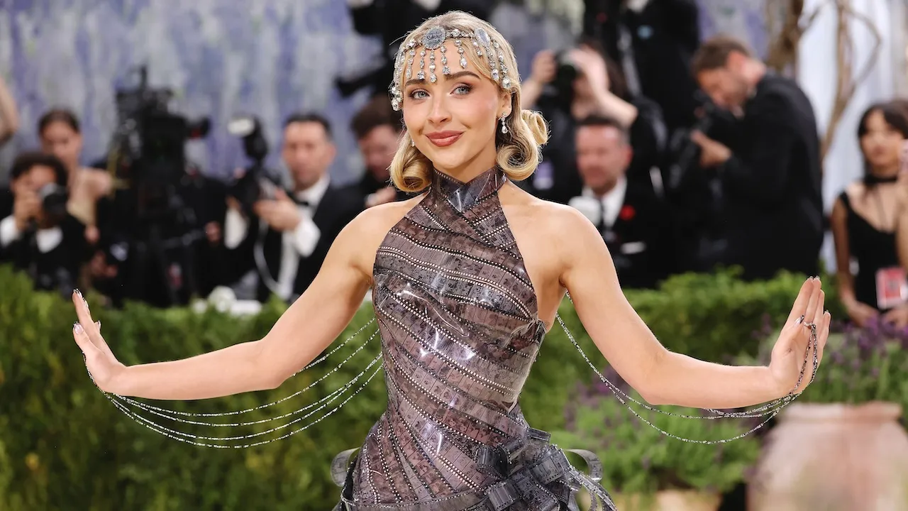 A woman wearing a sparkling metallic gown with crystal chain details and a jeweled headpiece poses on the Met Gala red carpet, with photographers and guests visible in the background.