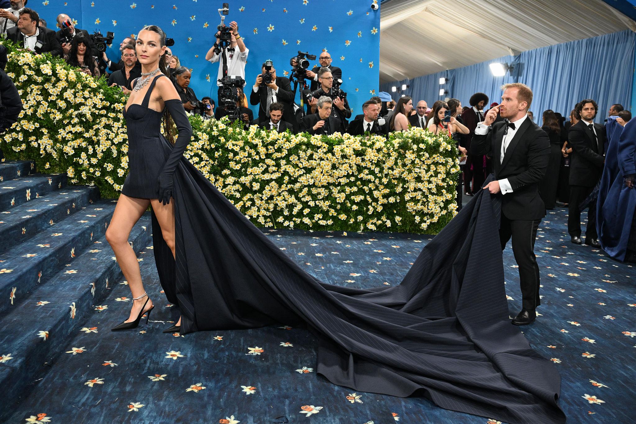 A woman wearing a fitted black gown with long gloves and an extended train poses on a floral-themed red carpet, while a man in a tuxedo carries the dramatic train behind her. Photographers and guests are visible in the background.